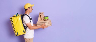 Side view portrait of friendly man in cap and thermo backpack, holding pizza boxes and takeaway coffee, handing order to client, copy space for promotional text, isolated on purple background.