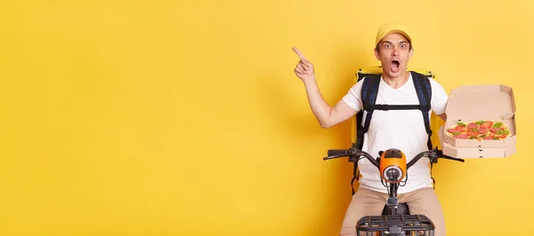 Indoor shot of shocked amazed courier man on bike holding open carton pizza box and pointing away at copy space for advertisement keeps mouth open, isolated over yellow background.