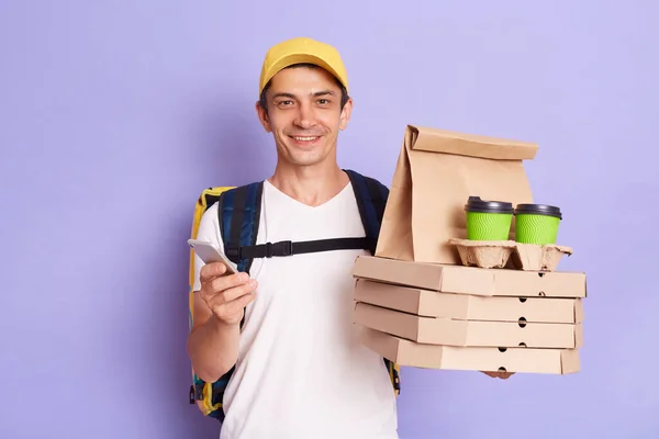 Image of happy delivery man in cap and white T-shirt using smart phone, holding pizza boxes and coffee, cheking address for handing parcel, isolated on purple background.