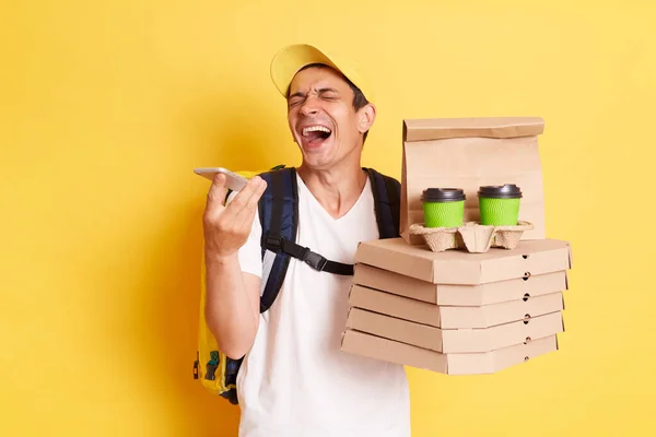 Image of extremely happy positive delivery man carrying parcel with pizza and coffee isolated over yellow background, holding mobile phone in hands, recording voice messages, laughing.