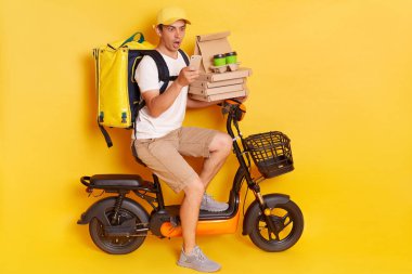 Shocked courier on electric scooter with thermo backpack delivering pizza and coffee to go, holding cell phone, looking at screen with open mouth, isolated over yellow background.