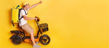 Shocked confident delivery man in cap riding bicycle with raised clenched fist, wearing thermo backpack bag isolated over yellow background. Mockup, advertisement area.