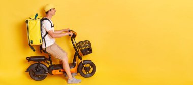 Fast and quick shipment service. Side view portrait of delivery man in cap riding bicycle, wearing thermo backpack bag, isolated over yellow background. Copy space for advertisement.