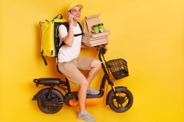 Kind friendly courier on electric bike with thermo backpack delivering pizza and coffee to go, talking on mobile phone and looking at camera with smile, isolated over yellow background.