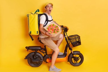 Portrait of astonished amazed courier on electric scooter with thermo backpack delivering pizza, looking at camera with big scared eyes fast food orders delivery posing isolated over yellow background