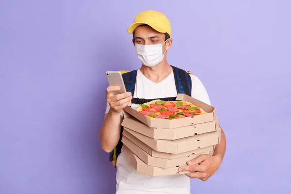 Fast service and best food delivery. Kind pleased male courier wearing protective mask, holding pizza boxes from restaurant and using smartphone, isolated on purple background.