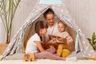 Horizontal shot of Caucasian mother with two daughters sitting together in teepee tent at home, playing with wooden eco toy, expressing positive emotions and happiness.