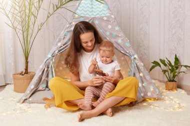 Indoor shot of attractive dark haired woman wearing white t shirt sitting in peetee tent with her infant baby daughter and playing with eco wooden toy, mother spending time at home with her kid.