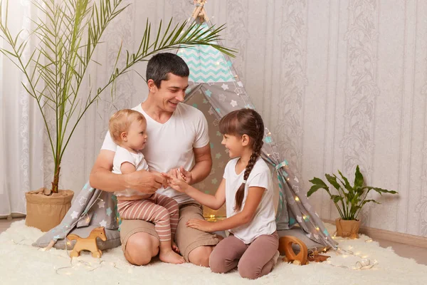 Indoor shot of smiling brunette man wearing white t shirt sitting in wigwam with his daughters and having fun, family playing together in peetee tent.