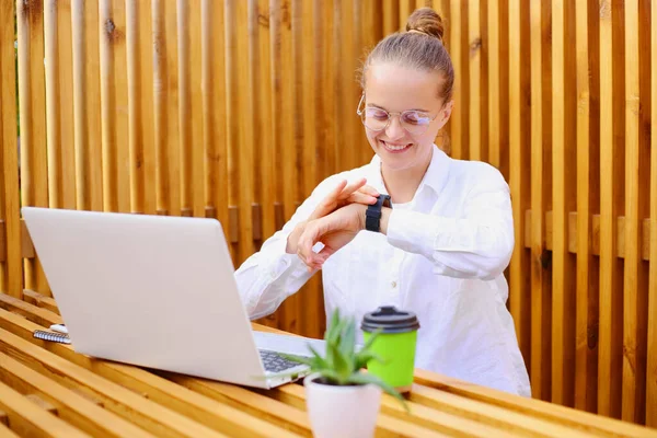 Young Caucasian business woman checking her watch while working on her laptop outdoors in the city cafe with wooden walls or outside office. Smiling happy female at the workplace.