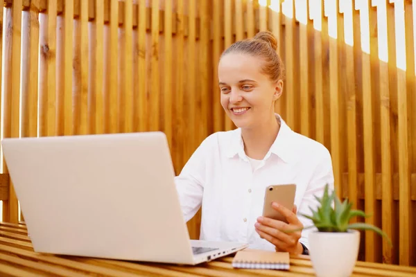 Horizontal shot of young adult woman with smart phone working on notebook, female wearing white shirt and glasses, sitting in office, expressing happiness, being busy at work.