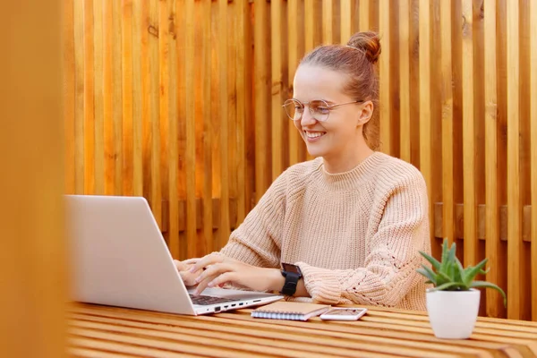 Portrait of smiling satisfied delighted woman wearing beige sweater working on laptop in office against wooden wall, typing on notebook with happy facial expression.