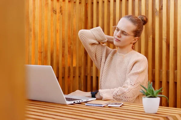 Image of tired exhausted woman wearing beige sweater working on laptop in office against wooden wall and feels pain in neck, works during long hours, massaging her back.