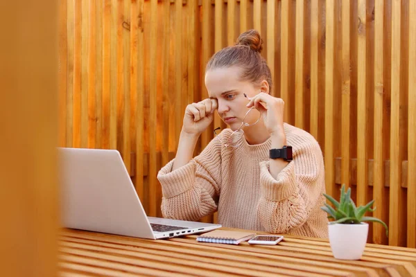 photo of exhausted woman wearing beige sweater working on notebook in office against wooden wall, rubbing her eyes, being tired, being sad and upset.