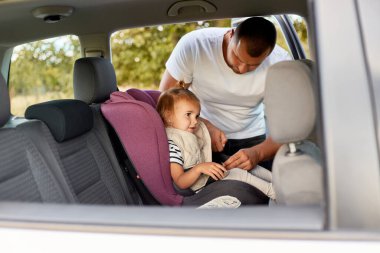 Portrait of young father wearing white t shirt securing cute baby with funny ponytail in the pink car seat in his car, children safety, preparing for a trip or travel.
