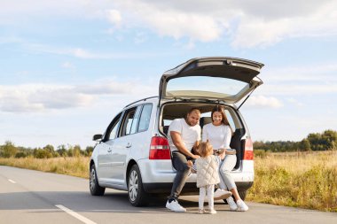 Outdoor shot of happy smiling woman and man drinking hot tea from thermos flask sitting in car trunk with their baby daughter, wife and husband spending good time together while traveling.