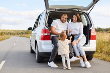 Horizontal shot of positive delighted family with infant kid drinking hot tea from thermos sitting in car trunk, stop their automobile for having rest.