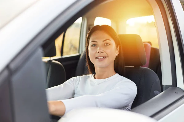 Portrait of happy woman driving a car and smiling, cute young success brunette woman driver steering car, wearing white shirt, looking at camera with satisfied expression.