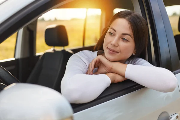 Horizontal shot of calm relaxed dark haired woman wearing white shirt sitting in the car and looking at the window, waiting for her passengers, posing during sunset.