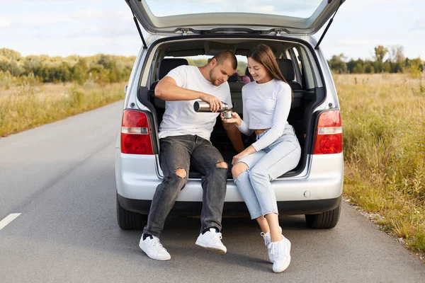 Tea party in car trunk. Portrait of loving couple drinks hot tea from thermos flask sitting in car trunk, family wearing white shirts and jeans traveling together, stop for rest and haning snack.