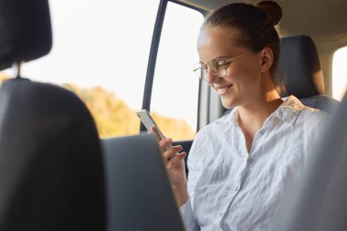 Gözlüklü, beyaz tişörtlü, bir arabanın arka koltuğunda otururken dizüstü bilgisayar ve cep telefonu kullanan, cep telefonundan mesaj okuyan ya da sosyal ağları kontrol eden bir kadın görüntüsü.