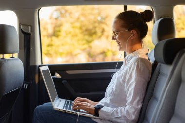 Gülümseyen beyaz bir iş kadınının portresi. Arabanın arka koltuğunda otururken taşınabilir bilgisayar kullanıyor. Bilgisayarda yazıyor..