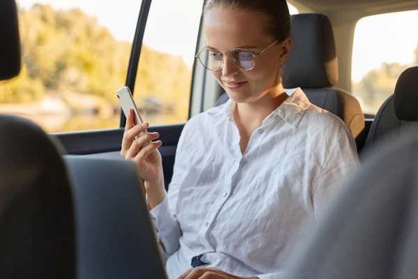 Horizontal shot of attractive businesswoman using notebook and smart phone for her online work while sitting on a backseat of a car, looking at display of laptop and holding mobile phone in hands.