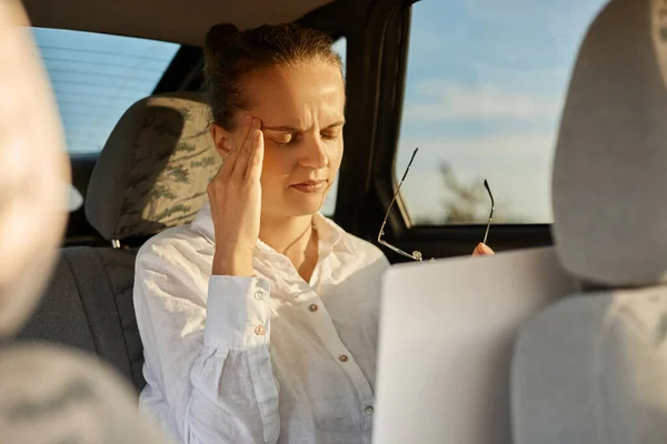 Portrait of sad depressed tired woman wearing white shirt sitting in car with laptop, being exhausted after hard working day, feeling pain in head, suffering headache, keeps eyes closed.