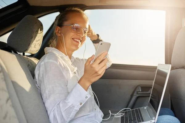 Profile portrait of adorable joyful businesswoman sitting on back seat of car and working with laptop, using smart phone for checking social networks or browsing internet while resting.