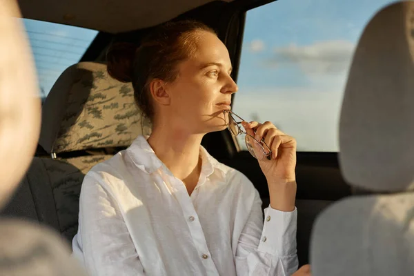Portrait of pensive calm relaxed attractive woman sitting on back seat in the car with glasses in hands, looking away at window, enjoying sunset, wearing white shirt.