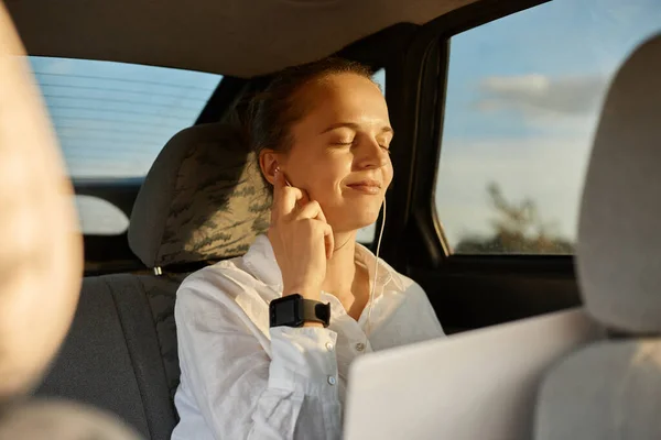 Photo of calm relaxed adorable woman wearing white shirt sitting on back seat of car and working with laptop, sits with closed eyes and enjoying music in her earphones.
