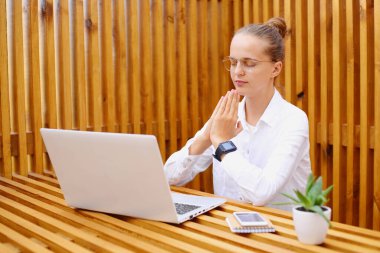 Beyaz tişörtlü, optik gözlüklü, beyaz saçlı, gevşek beyaz bir kadının laptopta oturup, yoga yaparken sakinleştiği ve konsantre olduğu bir fotoğraf..