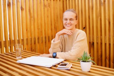 Beyaz saçlı, koyu renk saçlı, bej kazaklı, kapalı dizüstü bilgisayarın yanında oturan kadının portresi. İşini bitiriyor, mutlu görünüyor, kameraya bakıyor..