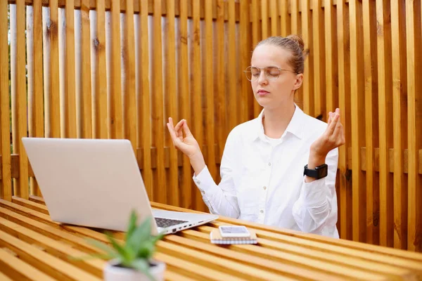 Portrait of calm young adult dark haired woman with hair bun wearing white shirt sitting and working on laptop, trying to calm down, meditating, practicing yoga during break.