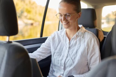 Saçlı, beyaz tişörtlü, bir arabanın arka koltuğunda otururken dizüstü bilgisayar kullanan, internette çalışan, gülümseyen, videolu bir kadın..