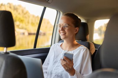 Gözlüklü güzel bir iş kadınının bir arabanın arka koltuğunda otururken dizüstü bilgisayar ve cep telefonu kullanması, sosyal ağları kontrol etmesi ya da komik videolar izlemesi..