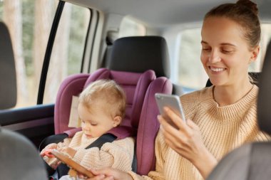 Mutlu, mutlu, mutlu bir annenin arabada kızıyla birlikte otururken telefonu kullanması. Kadın akıllı telefon ekranına bakıyor, sosyal ağları kontrol ediyor..