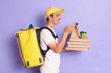Sarı şapkalı ve tişörtlü teslimatçının portresi termo sırt çantalı, karton kutuda pizza tutan ve menekşe arka planında izole edilmiş kahve alan cep telefonlu..
