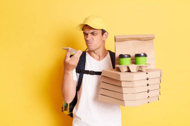Sarı şapkalı ve tişörtlü, elinde pizza ve kağıt bardaklarla müşteriyle iletişim kurmak için cep telefonu kullanan, sarı arka planda izole edilmiş bir kurye resmi..