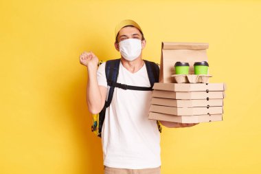 Sarı şapkalı teslimatçı, beyaz tişört ve koruyucu yüz maskesi elinde pizza ve kahve, sıkılmış yumruklar, neşeli, sarı arka planda izole edilmiş pozlar.