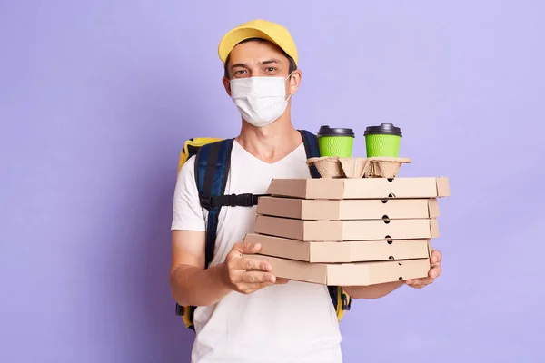 Indoor shot of delivery man in yellow cap, white t-shirt and protective face mask holding cardboard pizza and coffee, delivering food to client, posing isolated on purple background.