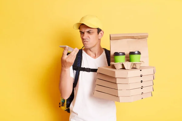 Image of angry courier man in yellow cap and t-shirt, holding pizza and paper cups of coffee, using cell hone for connecting with client, posing isolated over yellow background.