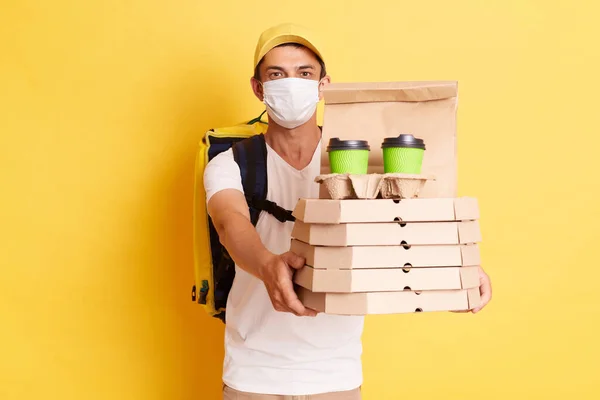 Portrait of delivery employee man in yellow cap, white t-shirt and protective face mask holding cardboard pizza and coffee, handing order to client, isolated on yellow background