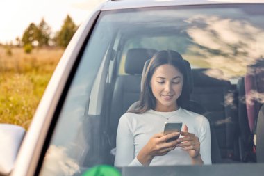 Arabada otururken cep telefonuna gülümseyen güzel, genç bir kadın. Çekici, koyu renk saçlı, beyaz tişörtlü, akıllı telefonuyla internette sörf yapan bir kadın..