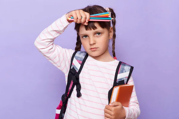 Horizontal shot of sick tired little girl in a striped shirt with notebook and pens, looking at camera with exhausted facial expression, suffering headache, posing isolated over lilac background.