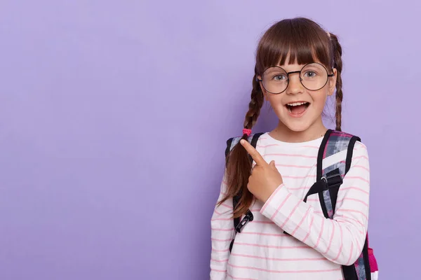 Horizontal shot of excited schoolgirl pointing with her fingers aside, showing copy space for promotional text, wearing striped shirt and glasses isolated on violet background.