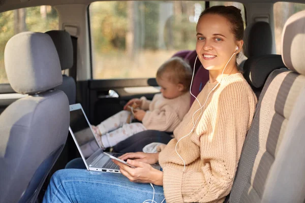 Side view portrait of delighted woman working on notebook while sitting with her baby daughter in safety chair on backseat of auto, looking at camera with happy expression, listening music.