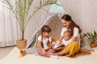 Anne ve iki sevimli küçük kızın çadırda yan yana oturduğu, anne ve çocuklarının çadır çadırında peri masalı kitaplar okuduğu kapalı bir fotoğraf..