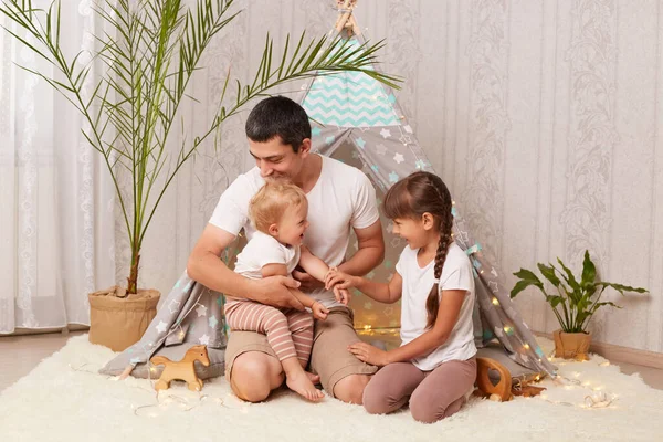 Indoor shot of attractive father and his little daughters having fun and playing in peetee tent in children room at home, playing with toys, spending joyful time together.
