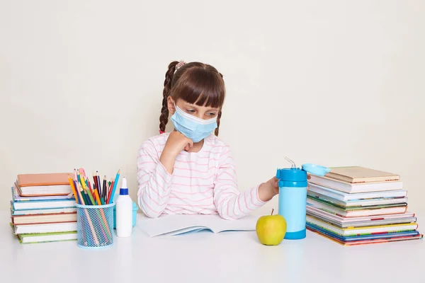 Indoor shot of cute sick unhealthy little schoolgirl wearing protective mask during visiting school, kid with dark hair and braids sitting at table surrounded with books, touching water bottle.